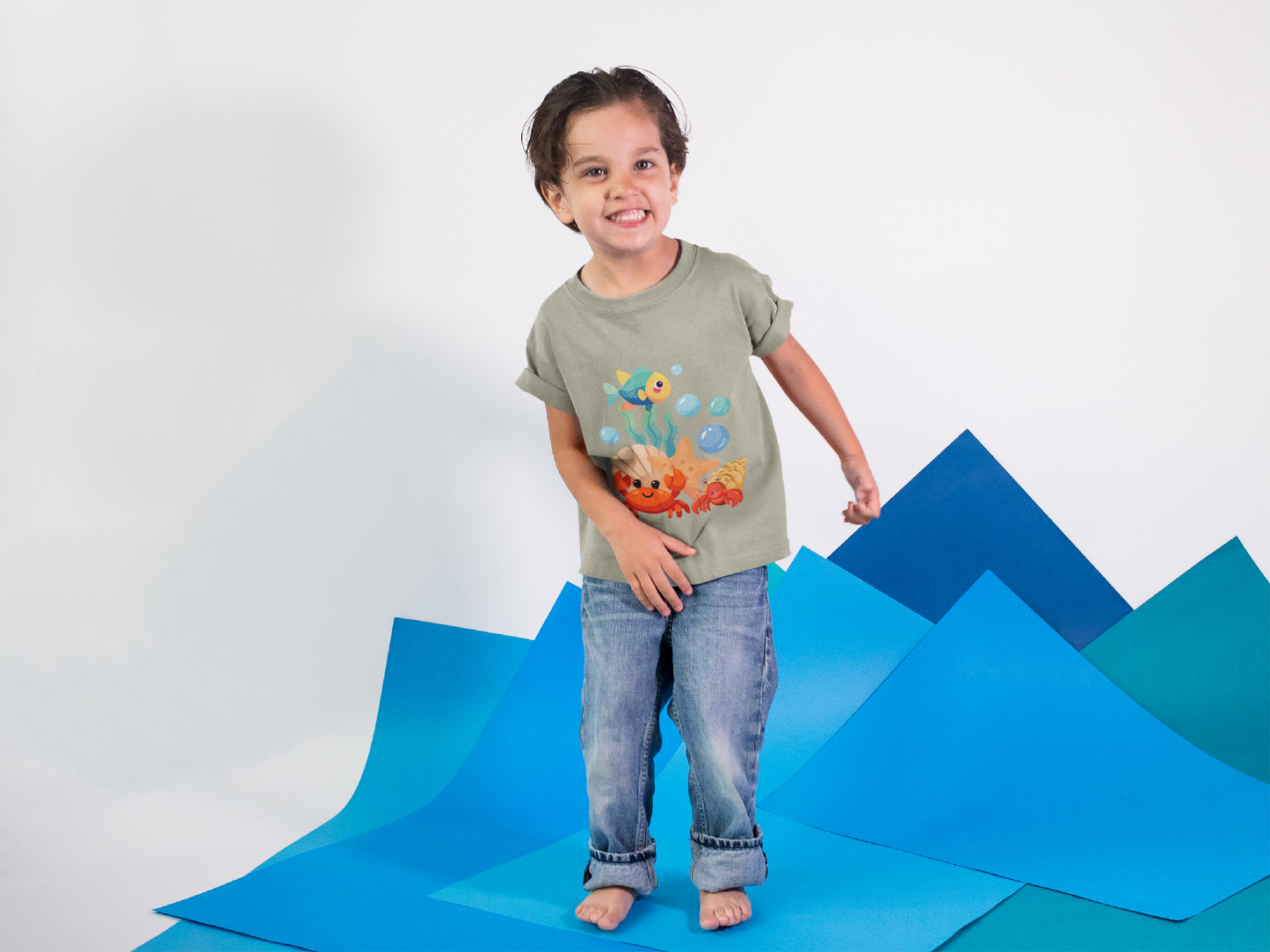 Child wearing a t-shirt with cartoon characters standing on a blue geometric surface. Playful toddler tee with a crab, fish & shell design. Soft, breathable cotton with tear-away label. Sizes 2T–5T. Perfect for beach days & summer play!