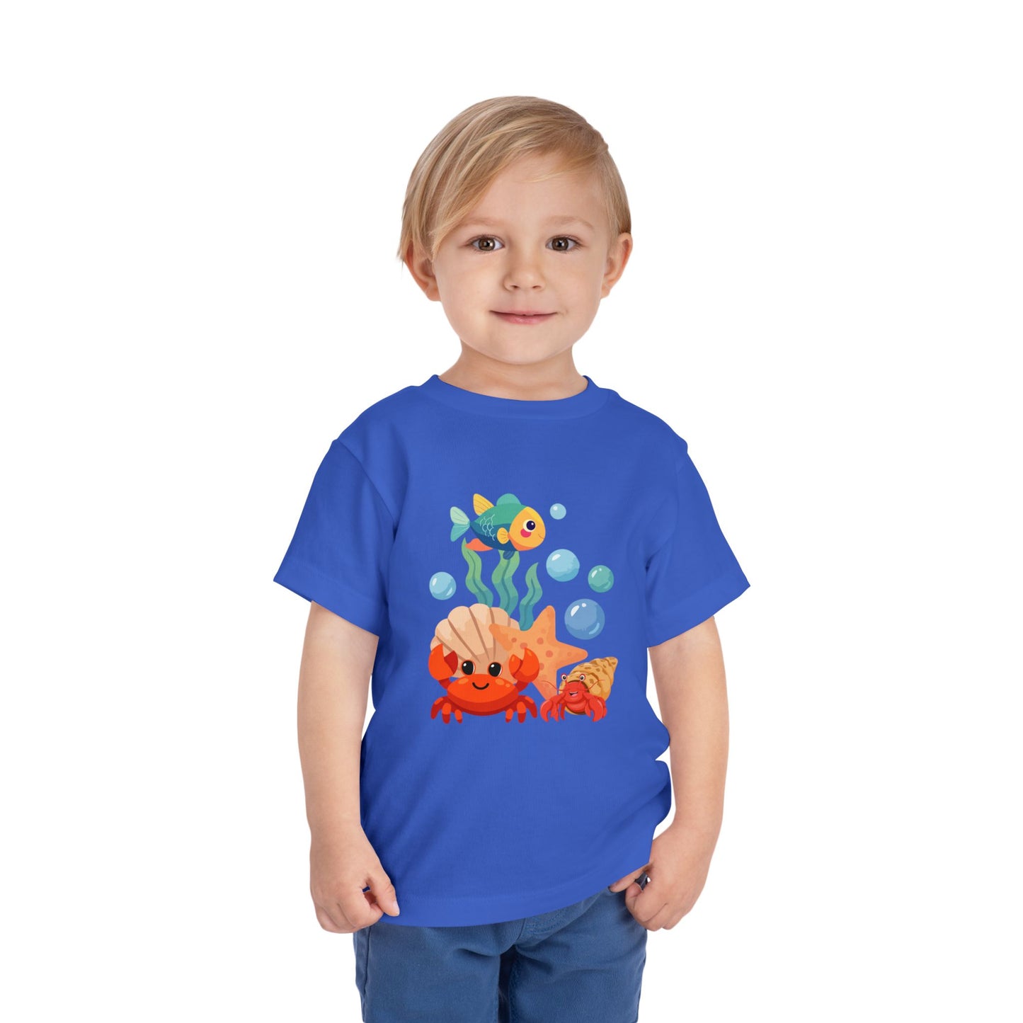 Child wearing a True Royal t-shirt with a colorful fish design on a white background. Playful toddler tee with a crab, fish & shell design. Soft, breathable cotton with tear-away label. Sizes 2T–5T. Perfect for beach days & summer play! 