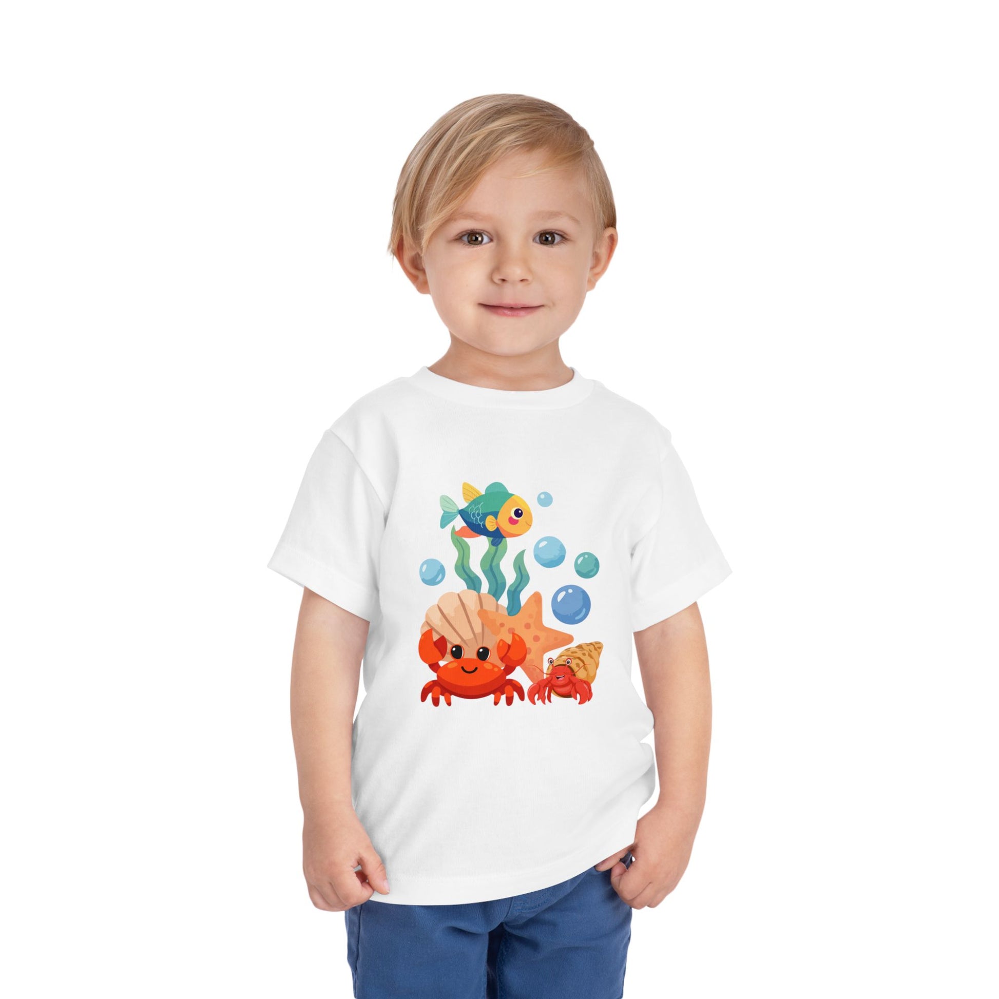 Child wearing a White t-shirt with a colorful fish design on a white background. Playful toddler tee with a crab, fish & shell design. Soft, breathable cotton with tear-away label. Sizes 2T–5T. Perfect for beach days & summer play!