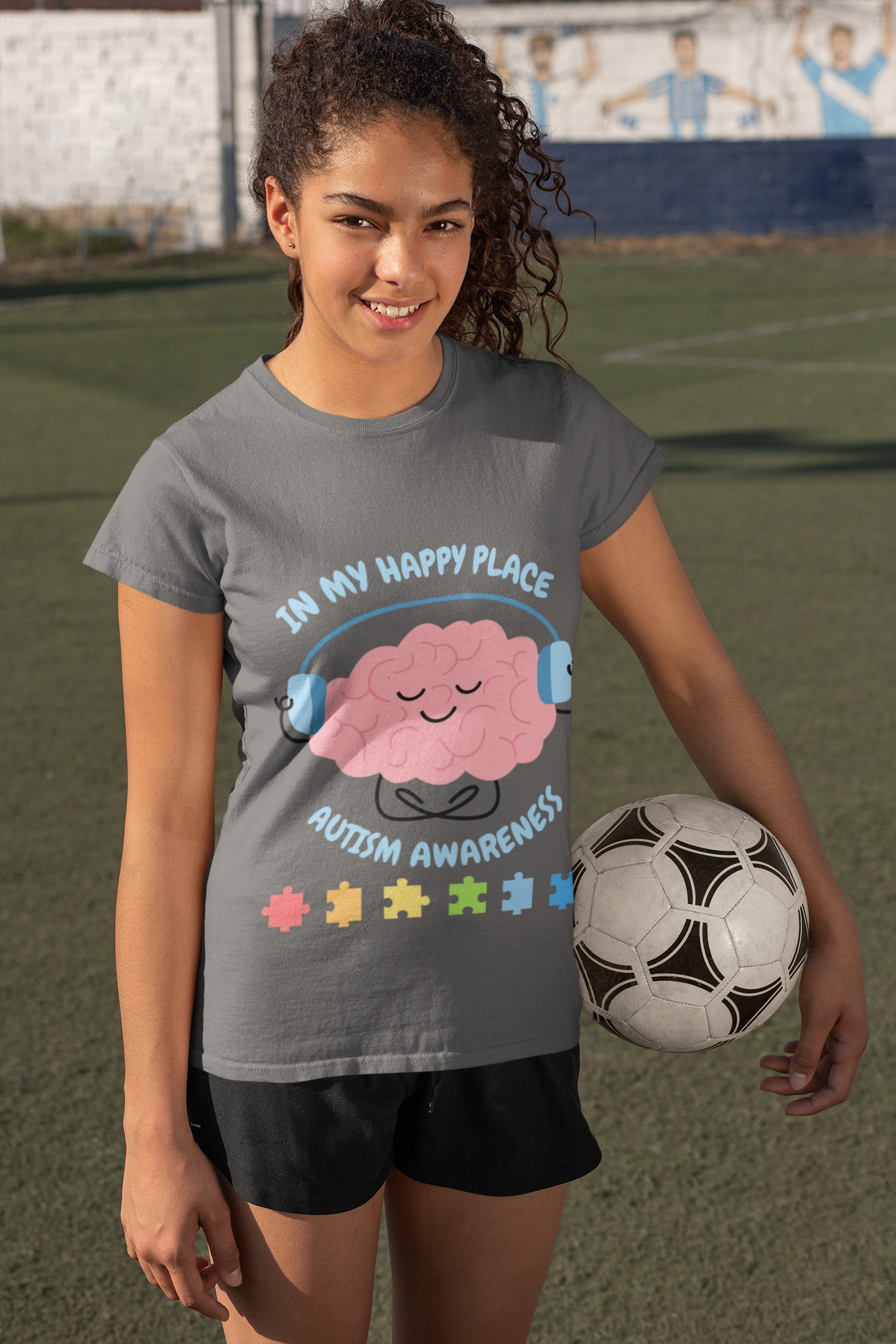 Girl wearing a t-shirt with an Autism awareness youth tee with smiling brain with headphones and colorful puzzle pieces, "In My Happy Place" graphic. Holding a soccer ball on a sports field. Asphalt