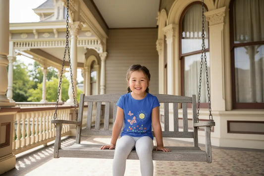 Toddler wearing this cheerful toddler tee with smiling sun, butterflies, and wildflowers, sitting on a swing of an old Victorian porch. Soft cotton, perfect for spring, playdates, and outdoor adventures. Bright kids' clothing. True Royal