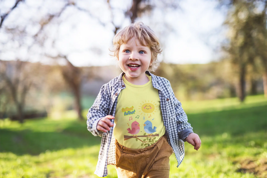 Child running outdoors in a park with trees and grass in the background.  Soft, lightweight toddler tee with cheerful singing birds & sunny design. Tear-away label, sizes 2T–5T. Perfect for playful days & gifting. Heather Yellow Gold